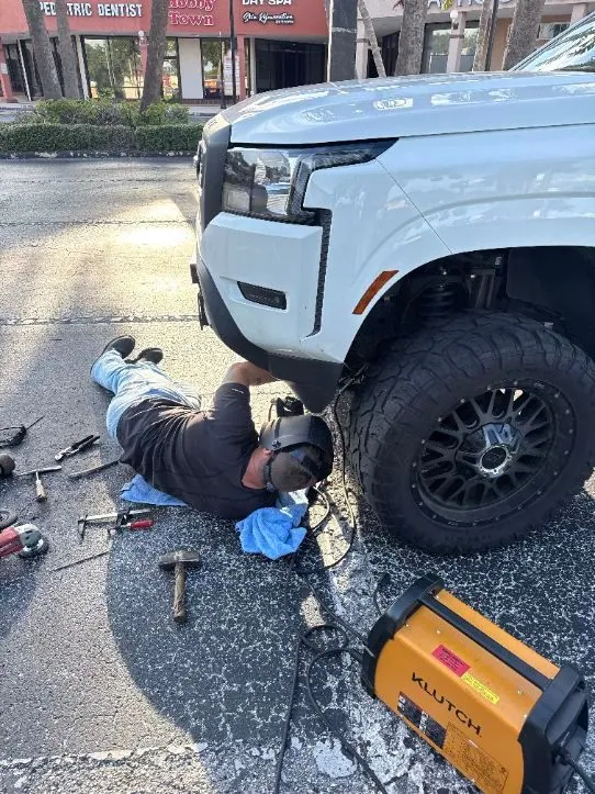 On-site welding under a lifted white pickup truck with a portable welder on the pavement