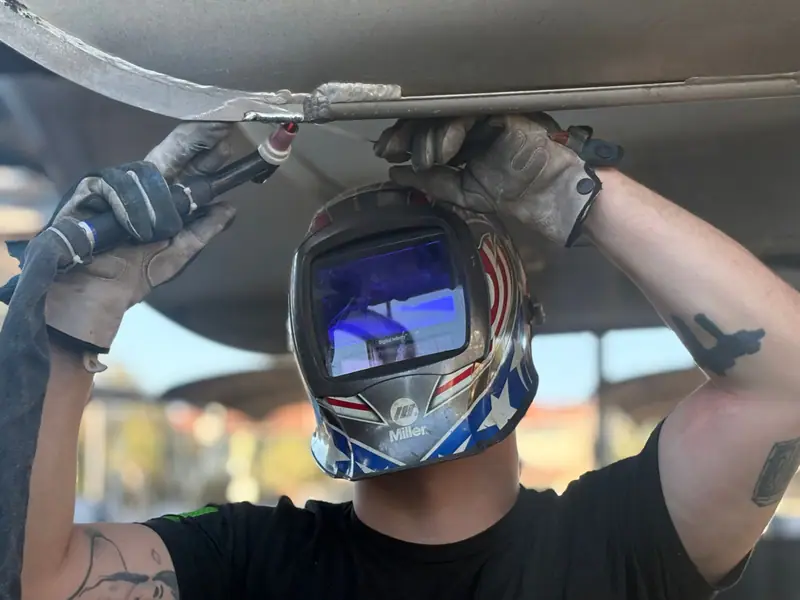 Welder running a TIG bead on the underside of a pontoon tube, filler rod in hand