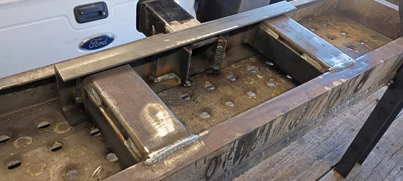 Welded steel bumper frame with corner brackets next to a white Ford Super Duty tailgate